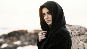 a person wearing a dark hoodie with the hood up as she stares this way, outdoor blurred background